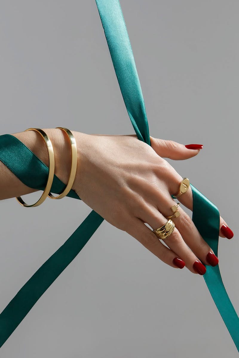 Close-up of elegant hands with gold jewelry and teal ribbon against a neutral background.